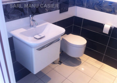 Meuble de salle de bain avec vasque blanche au dessus et toilettes à gauche. carrelage bleu foncé avec une frise